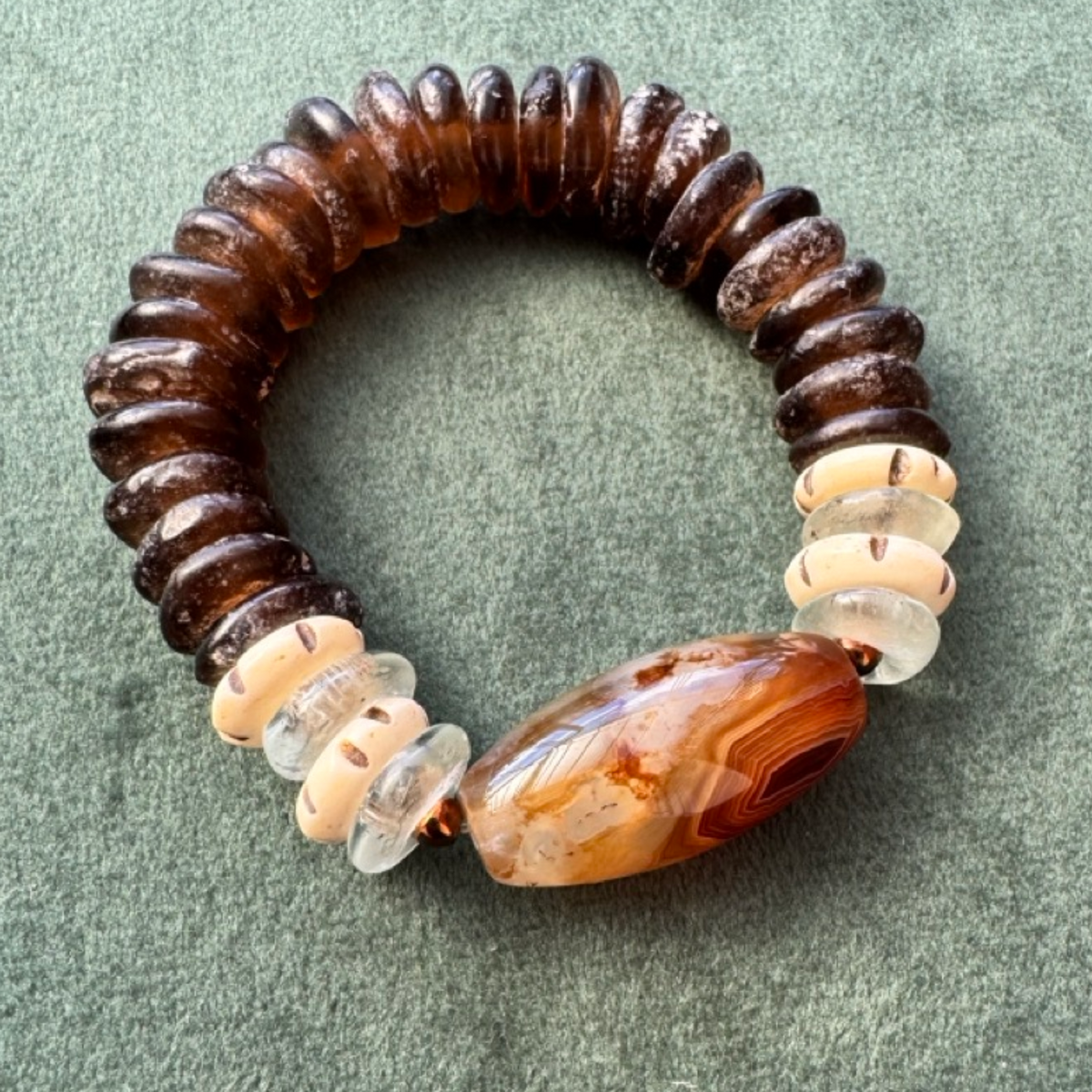 A handmade beaded stretch bracelet featuring dark brown agate beads, a large faceted agate bead with orange-brown banding, and clear crystal discs, displayed on a textured olive green background.
