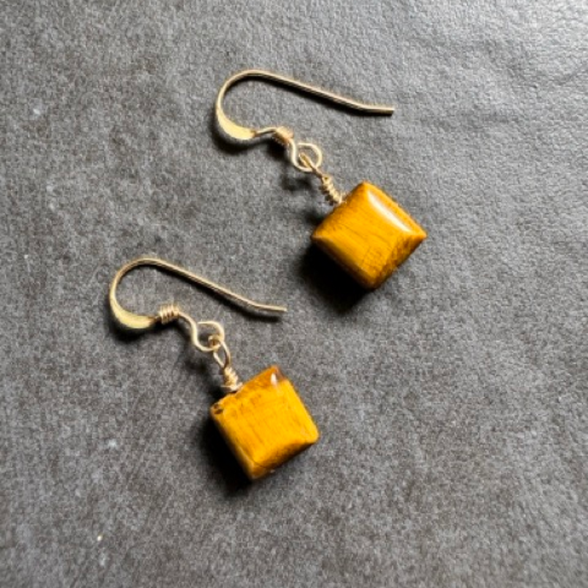 A pair of handmade dangle earrings featuring square yellow-gold tiger's eye stones on antiqued gold fishhook wires, displayed on a textured gray background.