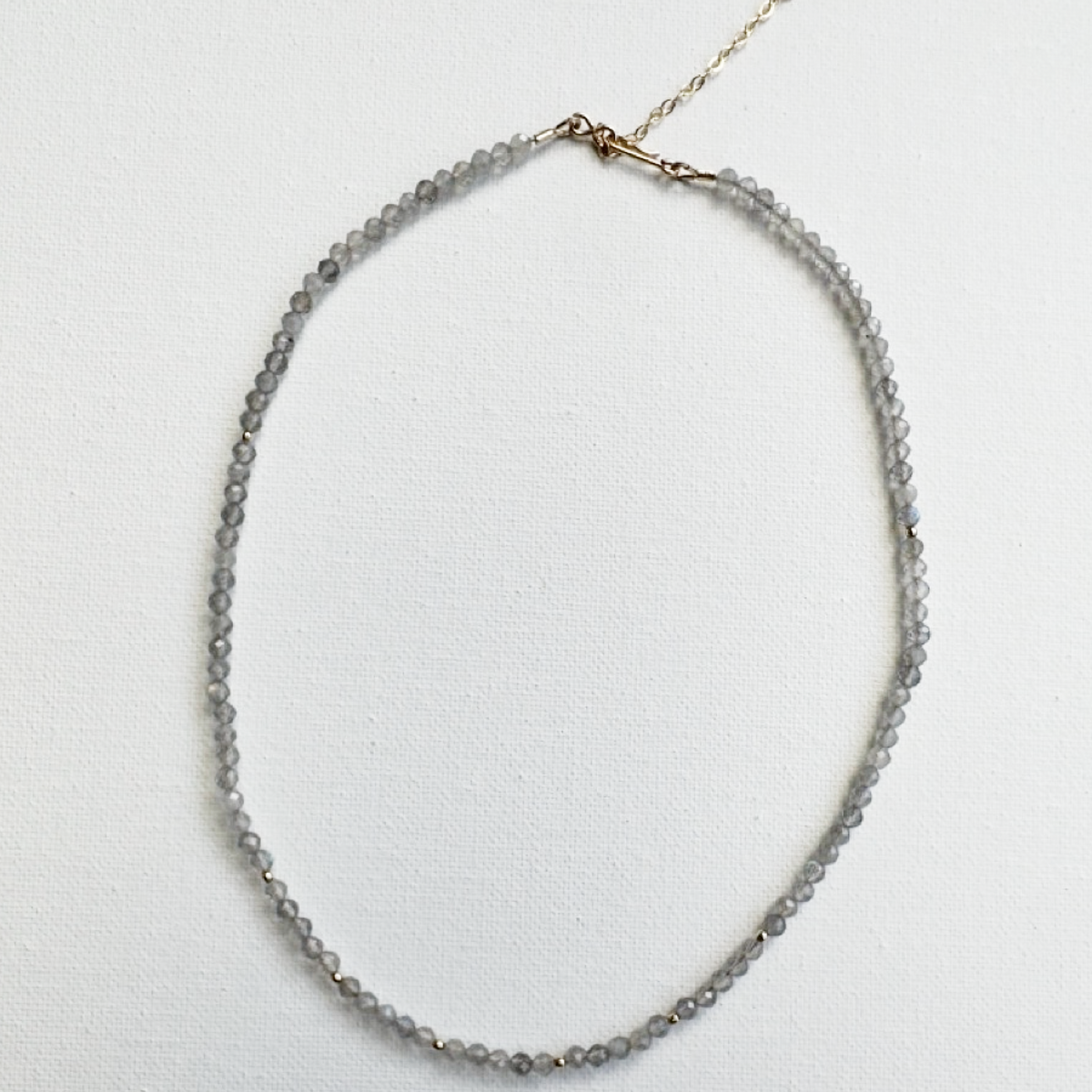 A delicate beaded necklace made of small gray faceted beads with a gold-tone clasp and chain, displayed on a textured white background.