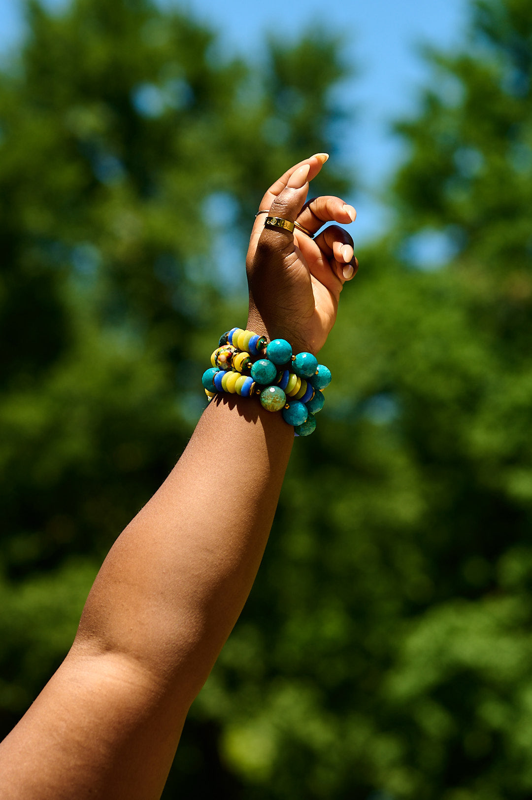 Woman's left arm raised in the air, wearing a stack of three blue, green, and yellow multicolored bead bracelets, in front of blurred background of green trees.