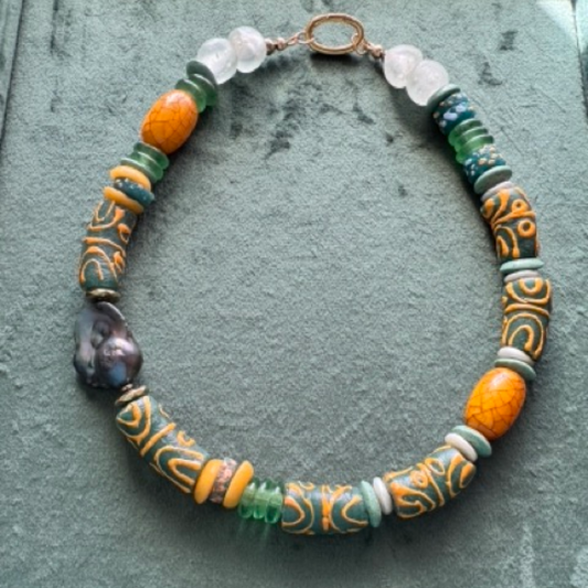 A handmade beaded necklace with a mix of patterned orange and green beads, a dark polished stone, and clear glass beads, displayed on a textured green background.