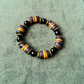 A beaded stretch bracelet featuring alternating black round beads and striped cylindrical beads in black, gray, and yellow, displayed on a textured green background.