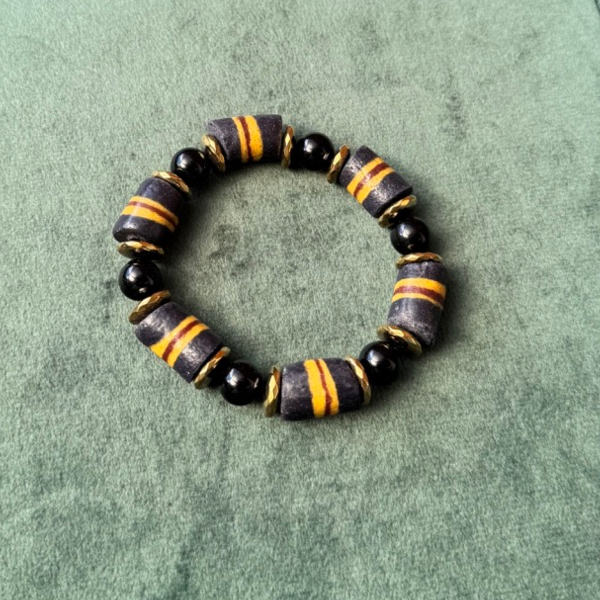 A beaded stretch bracelet featuring alternating black round beads and striped cylindrical beads in black, gray, and yellow, displayed on a textured green background.