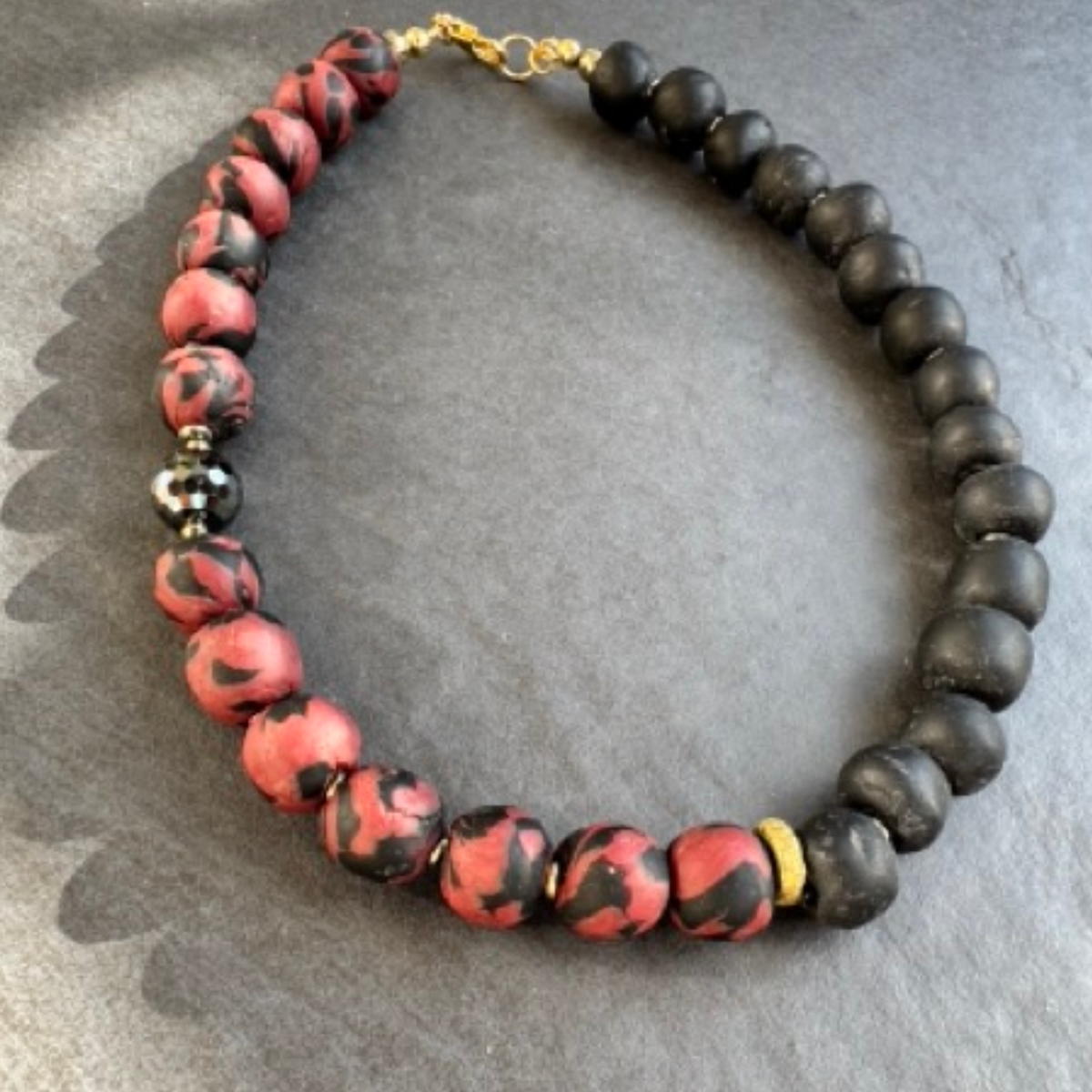 A beaded necklace with alternating matte black round beads and glossy red beads with black marbled swirls, accented by a faceted black bead and a tiny brass spacer, displayed on a gray textured surface.