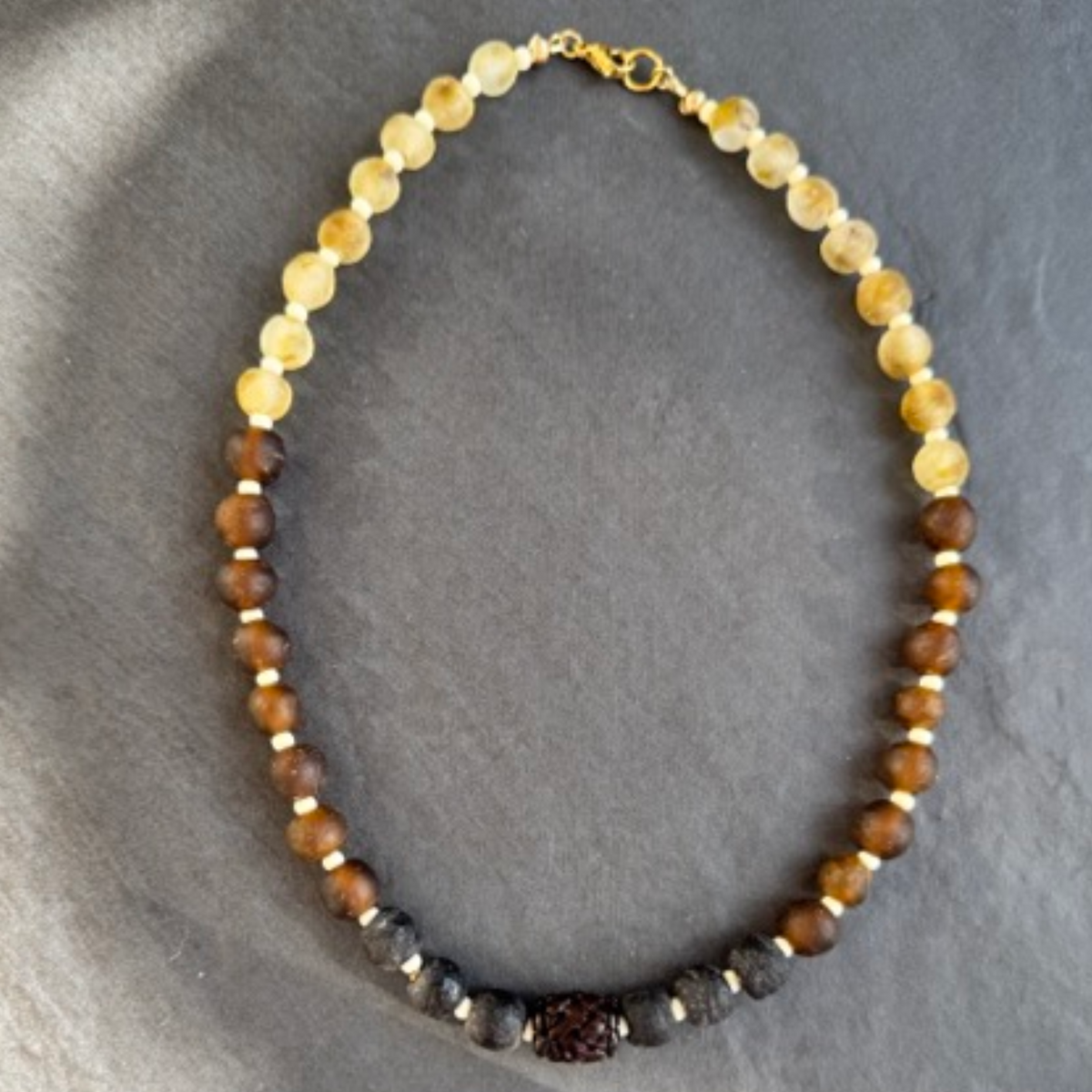 A beaded necklace made of alternating sections of translucent pale beige beads, medium brown beads, and dark charcoal black beads, with a small central dark brown bead and gold spacer accents, displayed on a dark gray slate background.