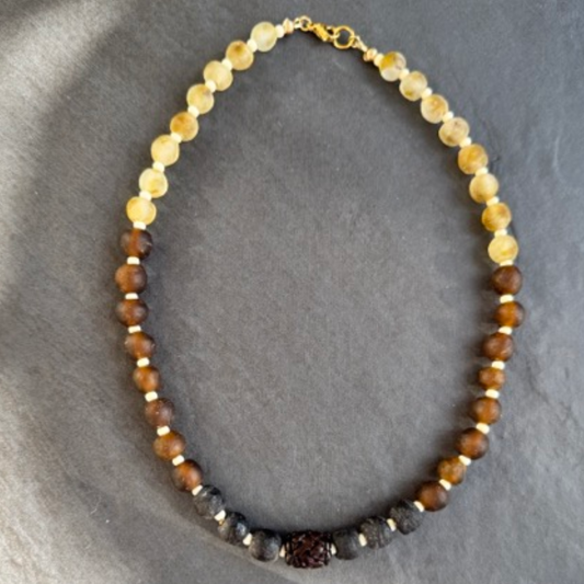 A beaded necklace made of alternating sections of translucent pale beige beads, medium brown beads, and dark charcoal black beads, with a small central dark brown bead and gold spacer accents, displayed on a dark gray slate background.