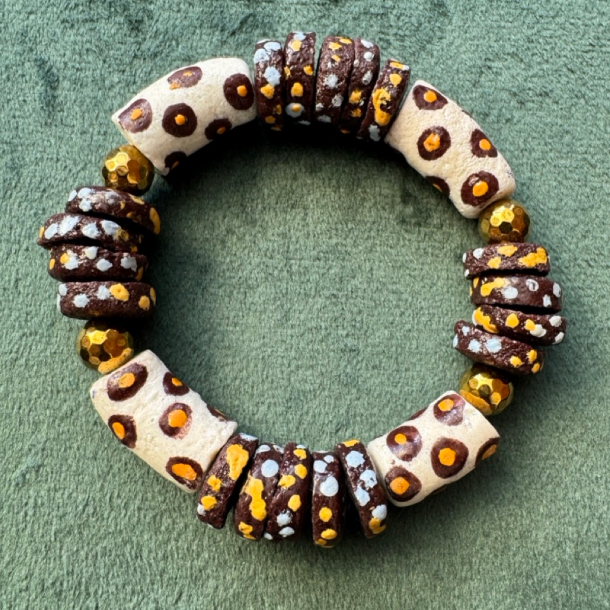 A handmade beaded bracelet made of alternating cream-colored beads with brown and orange leopard-like spots and dark brown beads with orange and white speckles, accented by faceted gold and white beads, displayed on a textured green background.