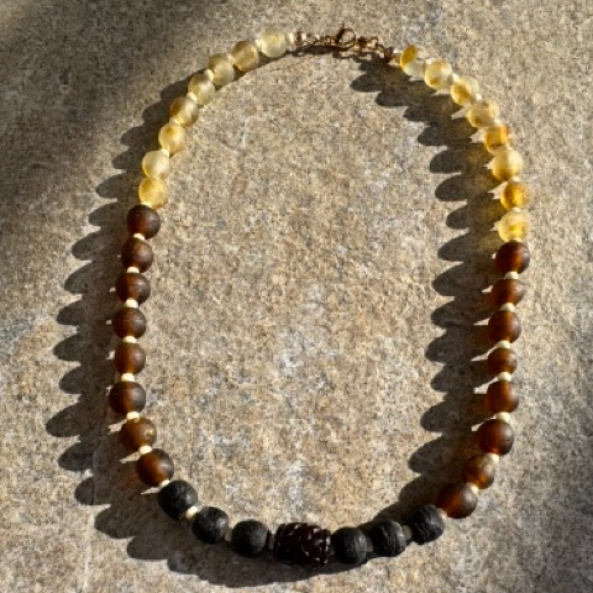 A beaded necklace made of translucent yellow beads, dark brown beads, matte black beads, and a central dark brown textured bead, displayed on a textured stone surface.
