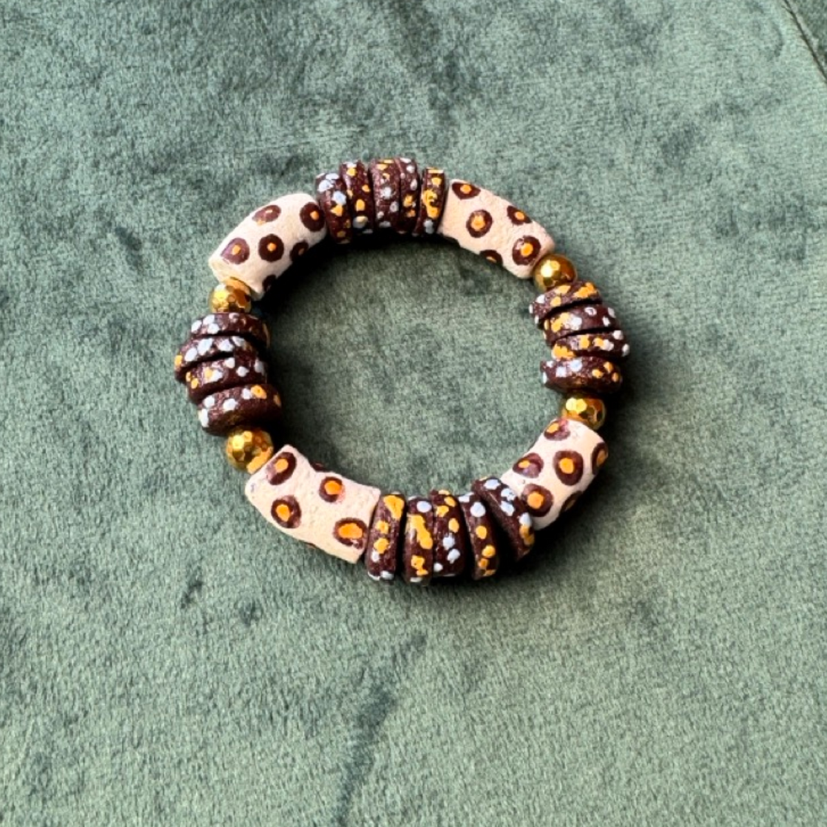 A handmade beaded bracelet made of alternating cream beads with brown and gold speckles and dark brown beads with white speckles, displayed on a textured green background.