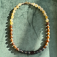 A beaded necklace with textured round beads in shades of amber brown, translucent pale beige, and dark charcoal black, arranged in an oval on a textured green background.