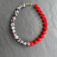 A beaded necklace arranged in an oval shape on a dark gray slate background. The bracelet features alternating sections of glossy red round beads and glossy black-and-white speckled beads, finished with a small gold clasp.