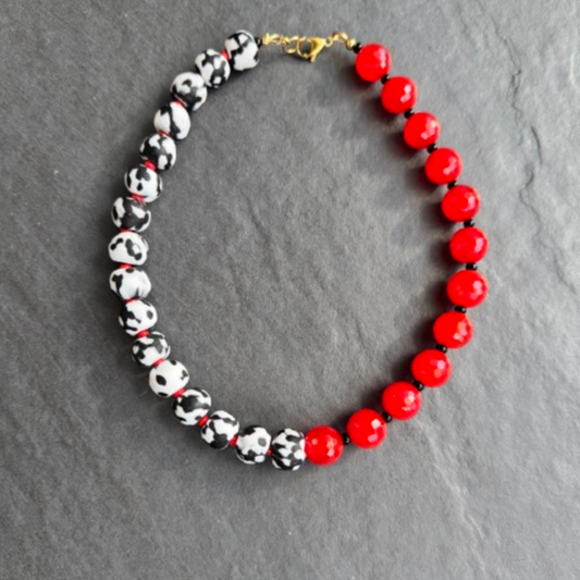 A beaded necklace arranged in an oval shape on a dark gray slate background. The bracelet features alternating sections of glossy red round beads and glossy black-and-white speckled beads, finished with a small gold clasp.