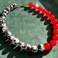 A beaded necklace with alternating black-and-white mottled beads and glossy red beads, finished with a gold clasp, displayed on a textured green surface.