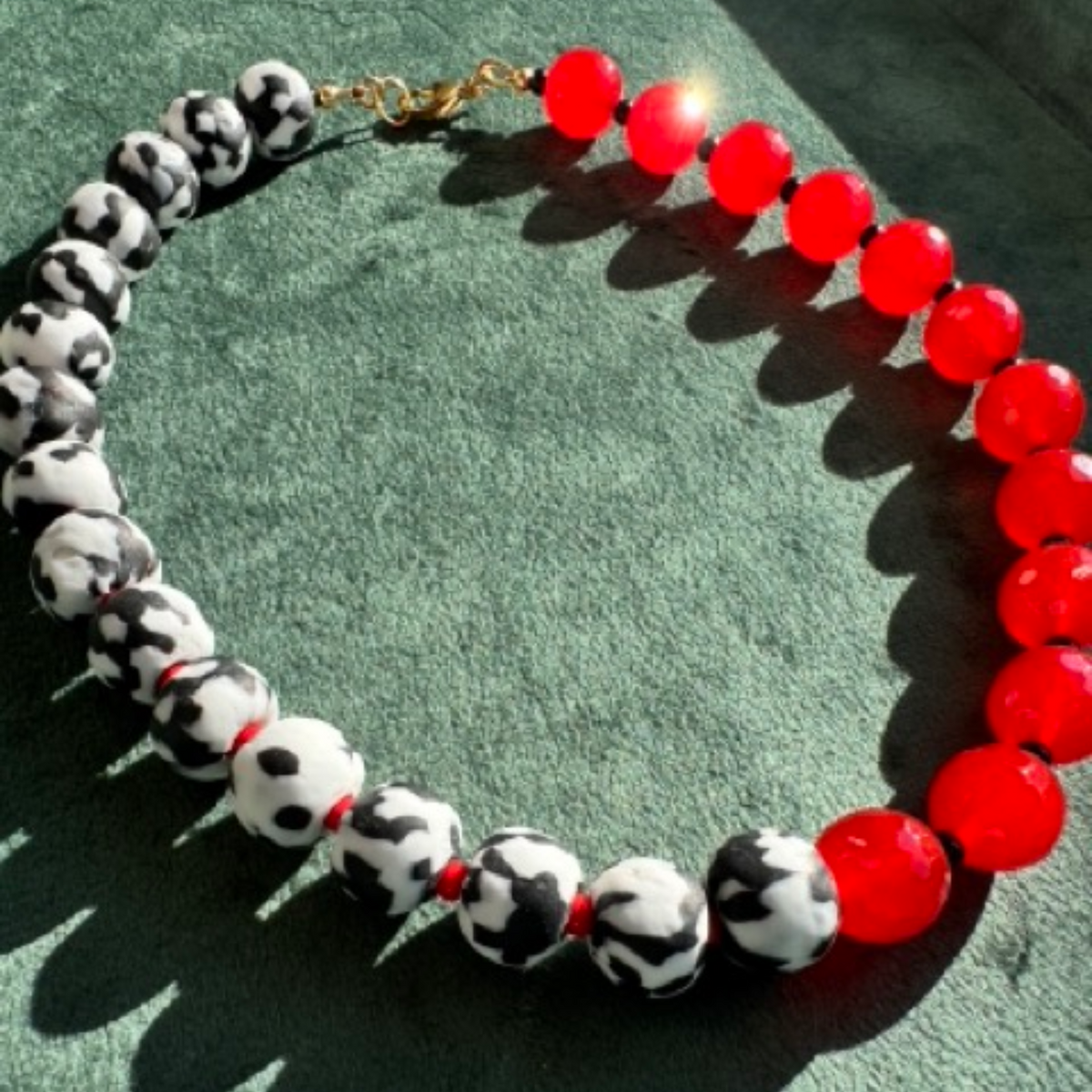 A beaded necklace with alternating black-and-white mottled beads and glossy red beads, finished with a gold clasp, displayed on a textured green surface.