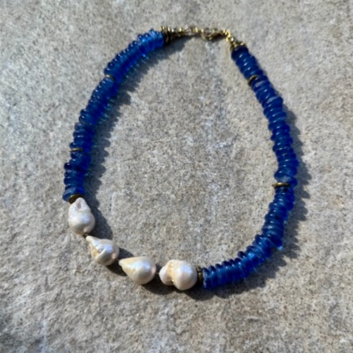 A handmade beaded necklace featuring dark blue glass beads, small gold spacer rings, and four baroque pearls, displayed on a textured gray surface.