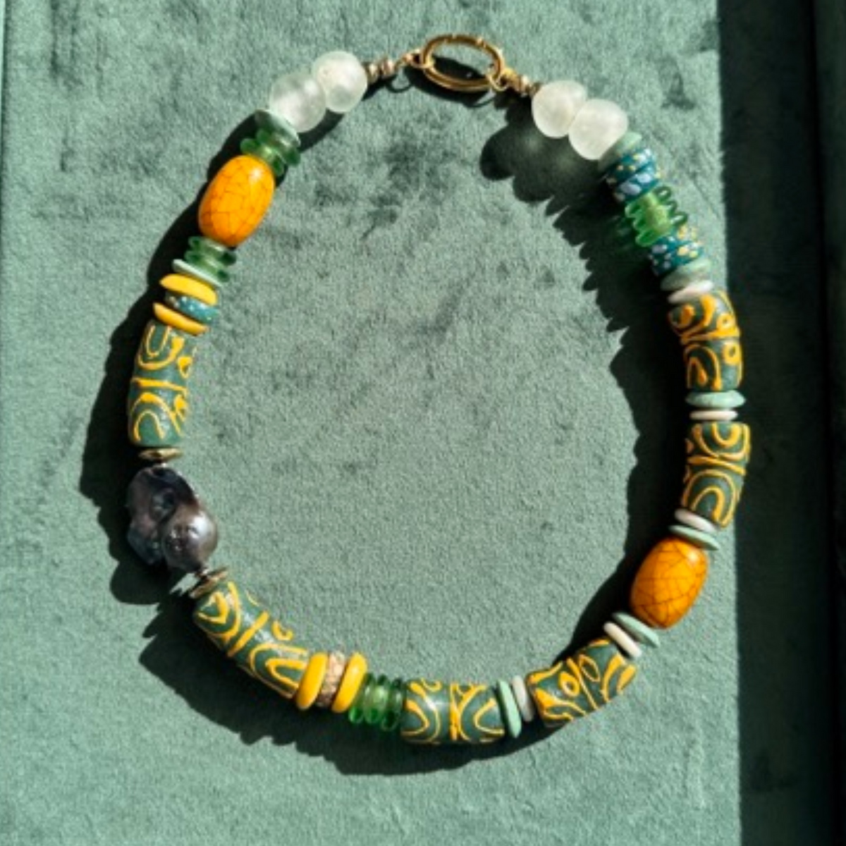 A beaded necklace with a mix of colorful cylindrical and round beads, including yellow, green, blue, white, and patterned yellow-and-blue beads, arranged in a circle on a textured teal background.