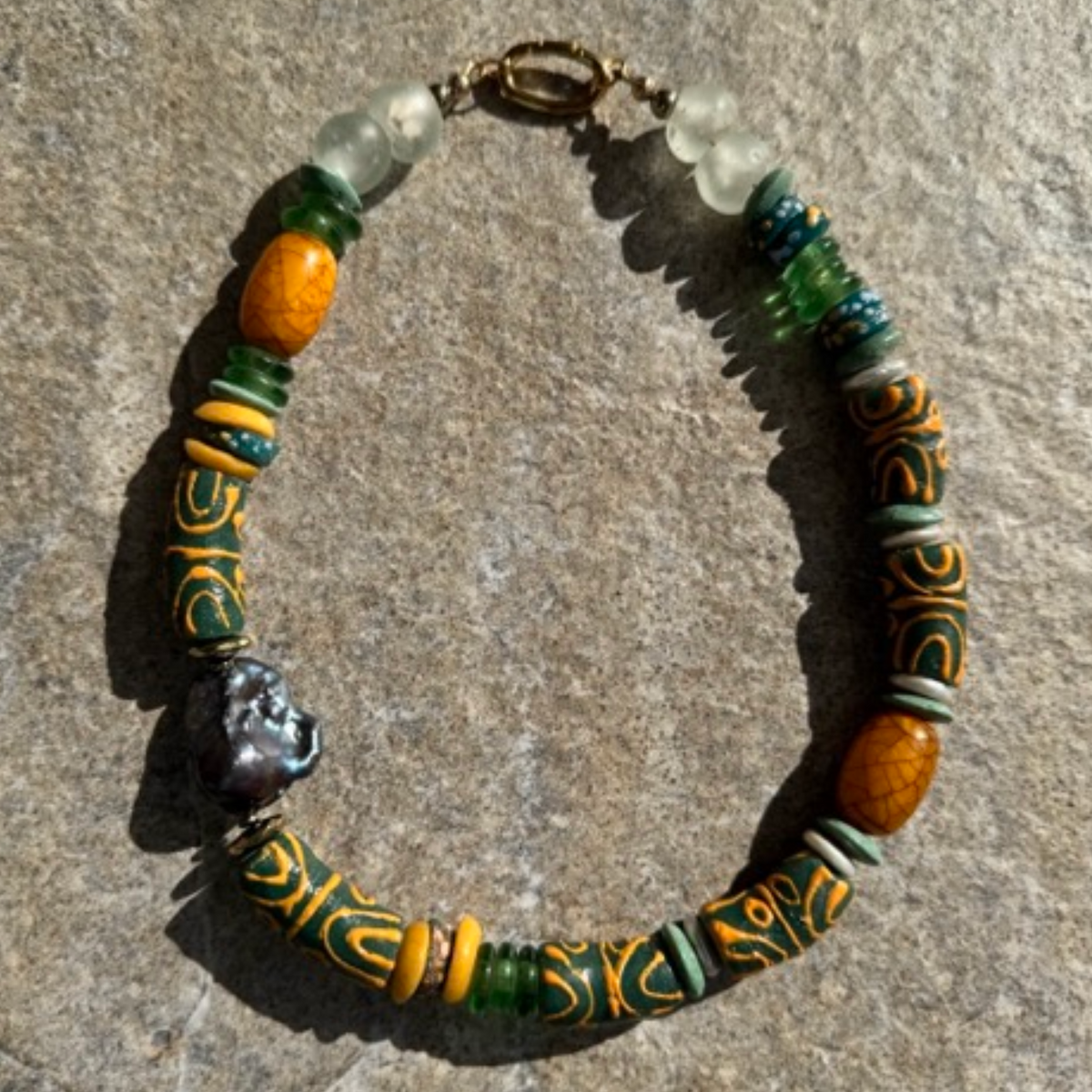 A handmade beaded necklace with a mix of patterned yellow and green beads, translucent white beads, a baroque pearl, and a gold clasp, displayed on a textured gray stone surface.