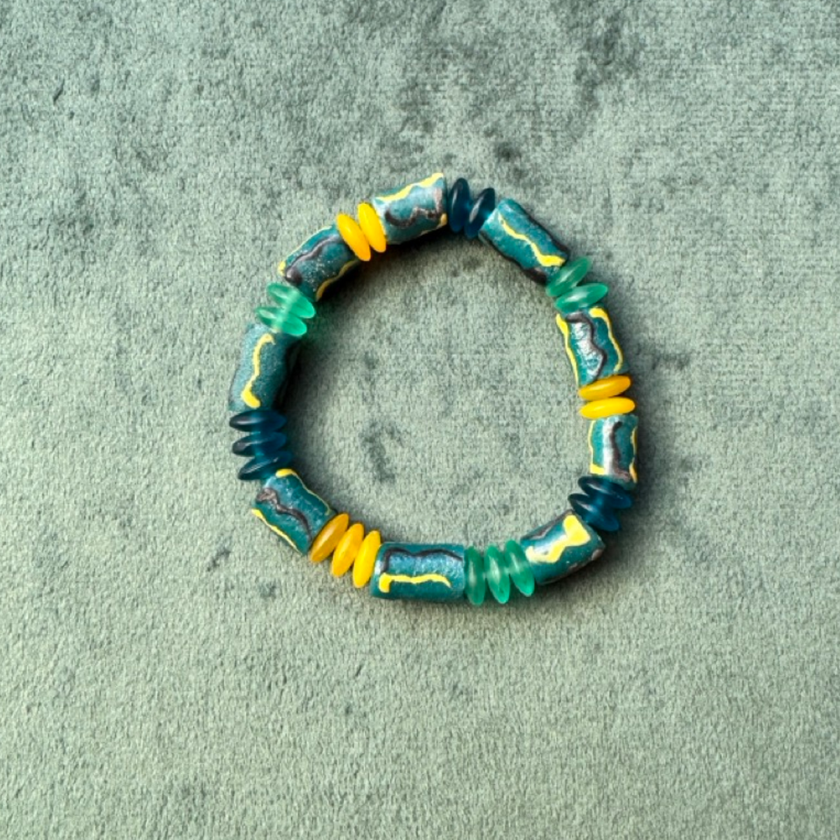 A handmade beaded stretch bracelet featuring textured turquoise, dark blue, and bright yellow beads, displayed on a textured gray-green fabric background.