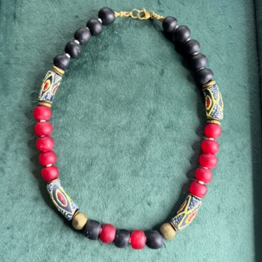A beaded necklace with alternating matte black and red round beads, accented by patterned cylindrical beads in black, yellow, and red, and small gold-toned spacer beads, displayed on a textured green background.
