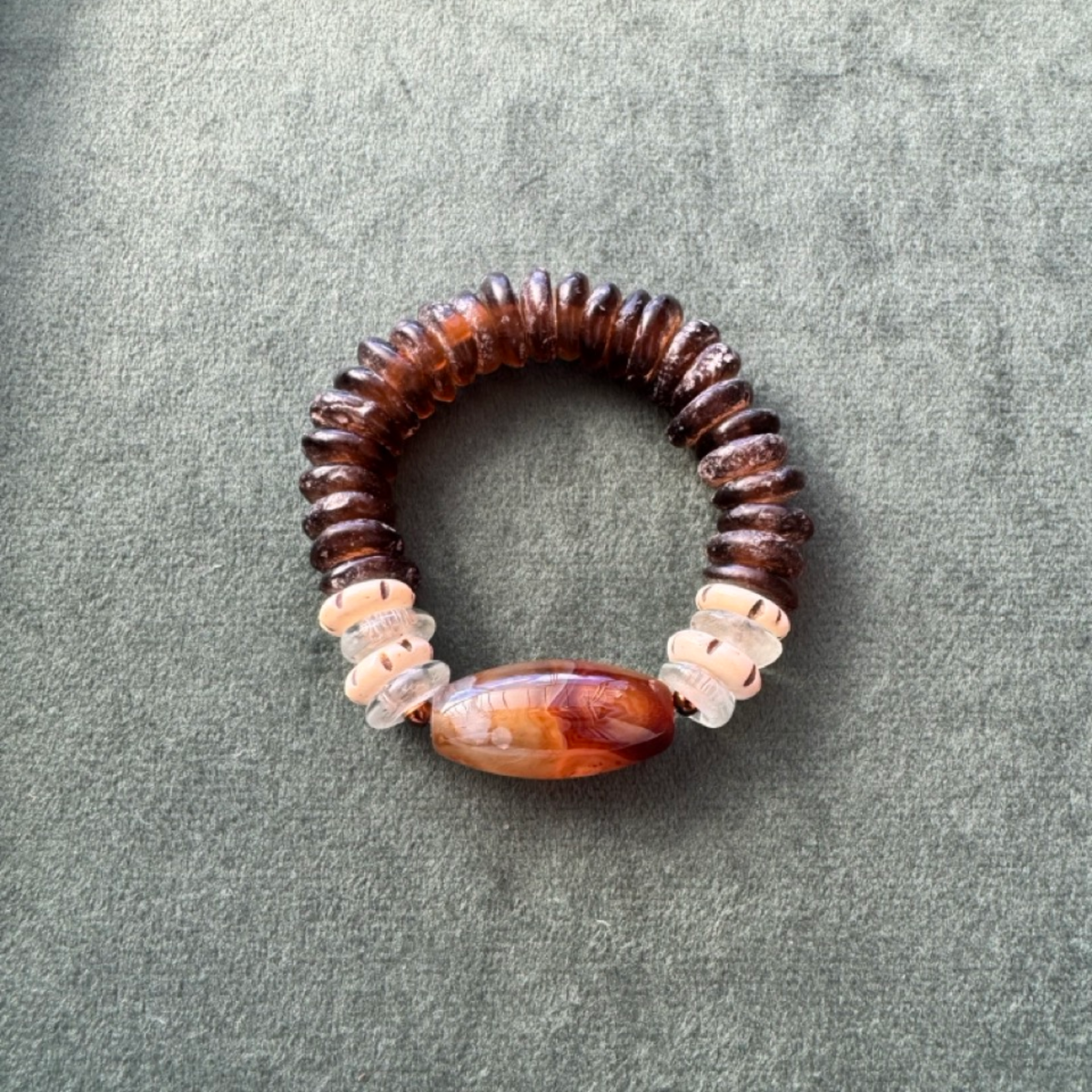 A handmade beaded stretch bracelet featuring dark brown disc beads, clear spacers, and a large polished oval agate bead in the center, displayed on a textured gray fabric background.