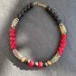 A beaded necklace with alternating black and red round beads, accented by patterned cylindrical beads in black, gold, red, and white, and two small brass beads, displayed on a dark gray slate background.