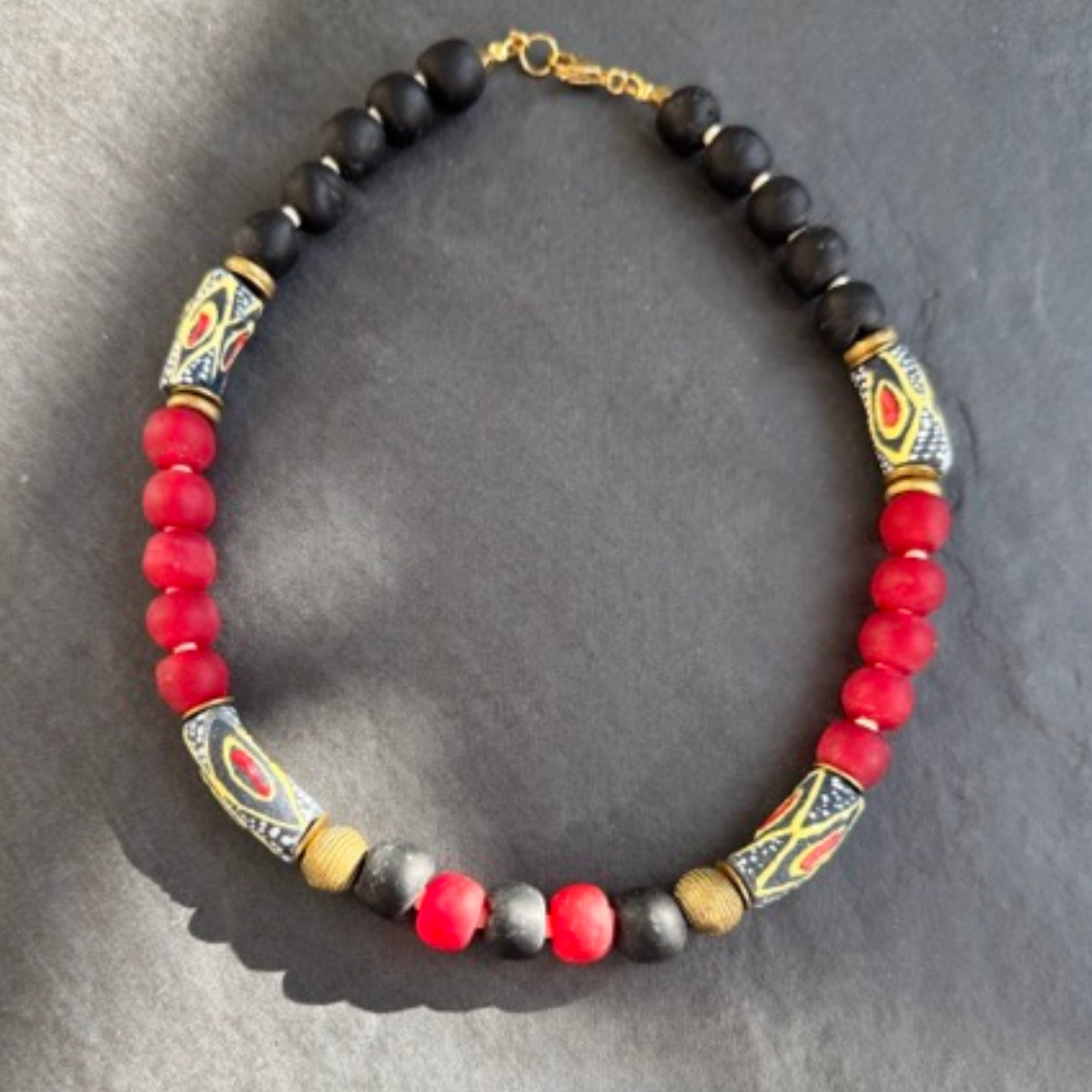 A beaded necklace with alternating black and red round beads, accented by patterned cylindrical beads in black, gold, red, and white, and two small brass beads, displayed on a dark gray slate background.