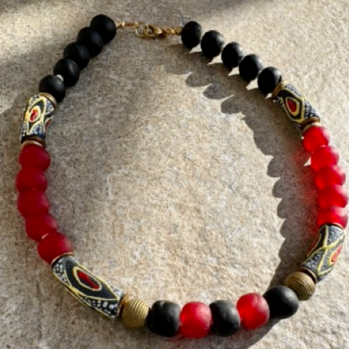 A beaded necklace with alternating matte black and translucent red beads, accented by patterned cylindrical beads in black, gold, and red, and a gold clasp, displayed on a textured beige stone surface.