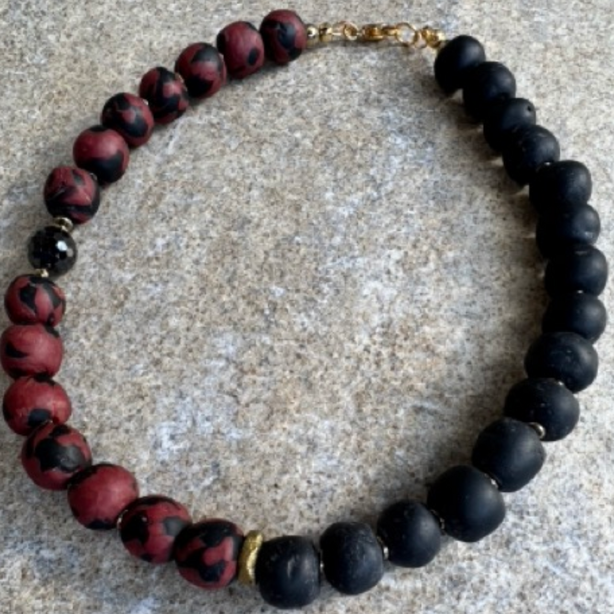 A handmade beaded necklace featuring alternating matte black and mottled red-and-black beads, with a small gold spacer and clasp, displayed on a textured gray stone surface.