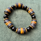 A beaded stretch bracelet featuring dark gray and black cylindrical beads with orange stripes, separated by small gold-toned spacer rings and accented with glossy black round beads, displayed on a textured green background.