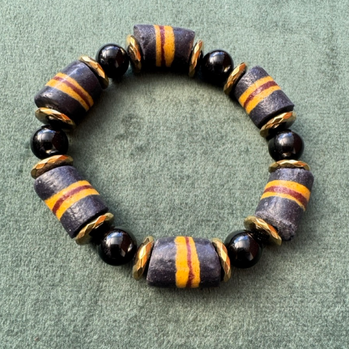 A beaded stretch bracelet featuring dark gray and black cylindrical beads with orange stripes, separated by small gold-toned spacer rings and accented with glossy black round beads, displayed on a textured green background.