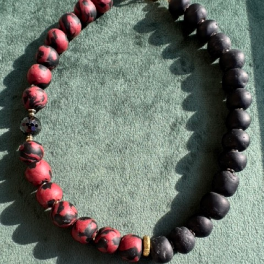 A beaded necklace with alternating matte black and red marbled beads, accented by a small faceted black bead and a tiny gold spacer, displayed on a textured green surface.