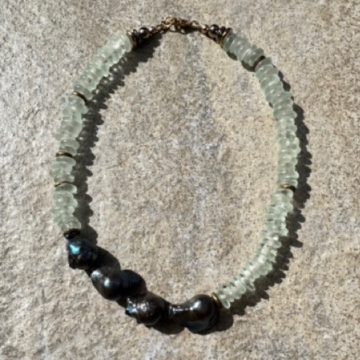 A beaded necklace featuring light green translucent stone beads, dark metallic baroque pearls, and small gold spacer rings, displayed on a textured gray stone surface.