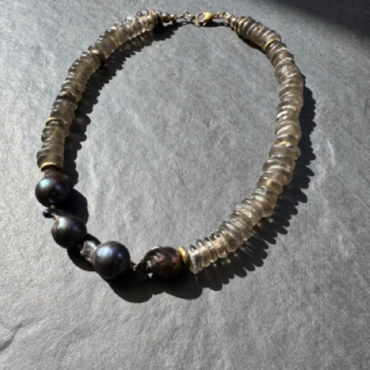 A beaded necklace made of translucent smoky gray beads with four dark iridescent pearls, displayed on a textured gray surface.