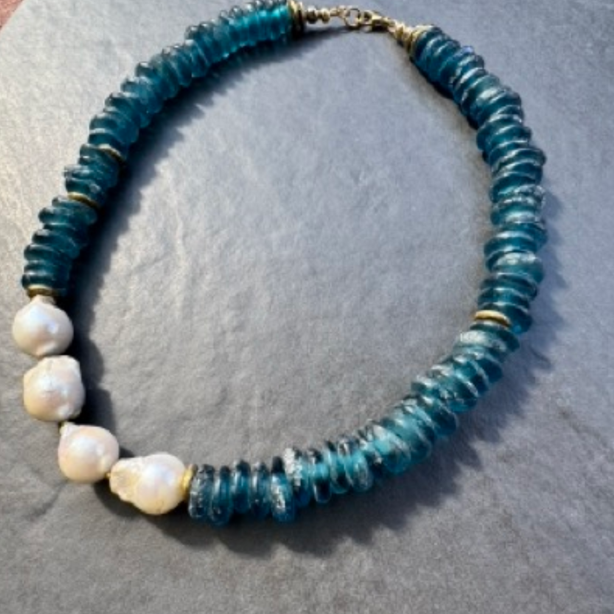 A handmade beaded necklace featuring textured teal-blue glass discs and four irregular white baroque pearls, accented with small gold spacer beads, displayed on a gray textured surface.