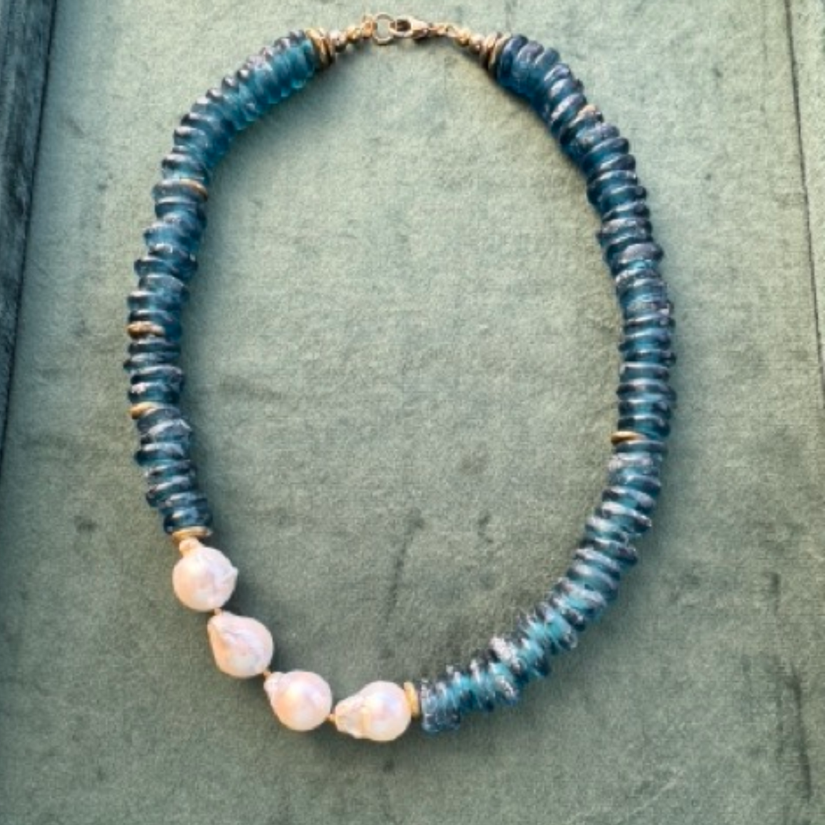 A handmade beaded necklace featuring textured blue-green glass beads and four irregular, pale pink baroque pearls, displayed on a textured green surface.