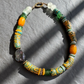 A handmade beaded necklace with a mix of green, orange, yellow, and white beads, including patterned cylindrical beads and a baroque pearl, displayed on a gray textured surface with diagonal shadows.