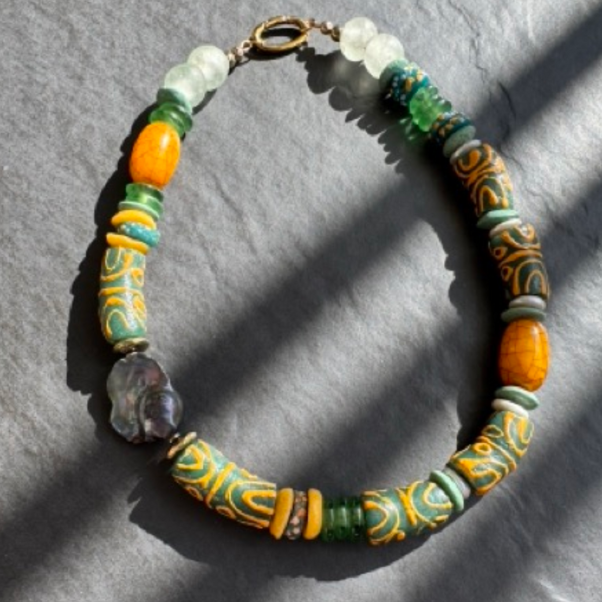 A handmade beaded necklace with a mix of green, orange, yellow, and white beads, including patterned cylindrical beads and a baroque pearl, displayed on a gray textured surface with diagonal shadows.