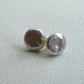 A pair of round pearl stud earrings with a lustrous silver-gray sheen, resting on a textured off-white fabric background.