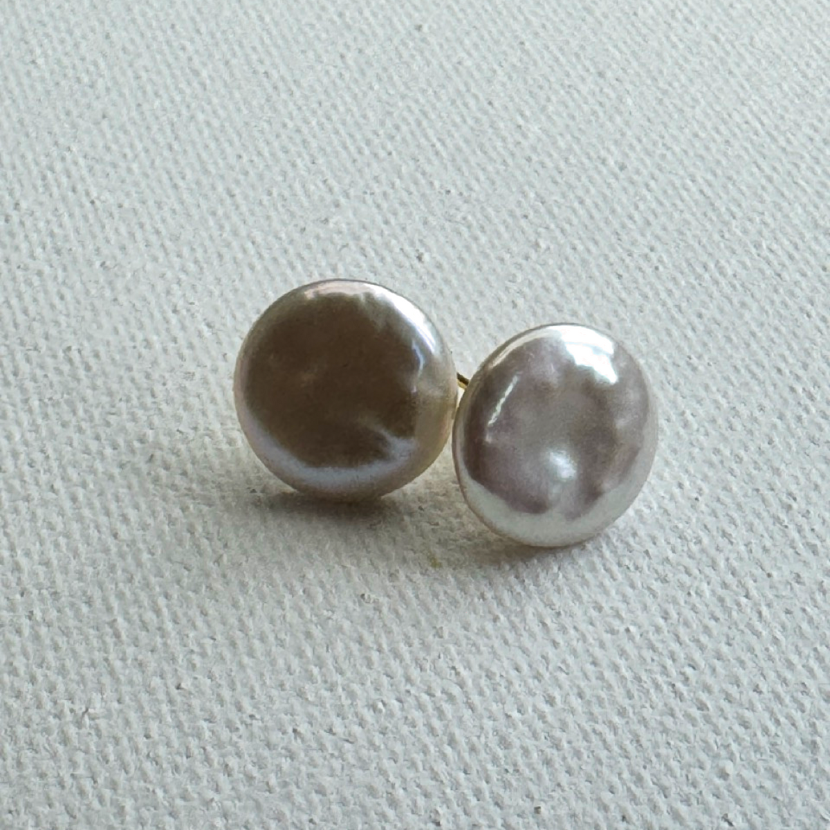 A pair of round pearl stud earrings with a lustrous silver-gray sheen, resting on a textured off-white fabric background.