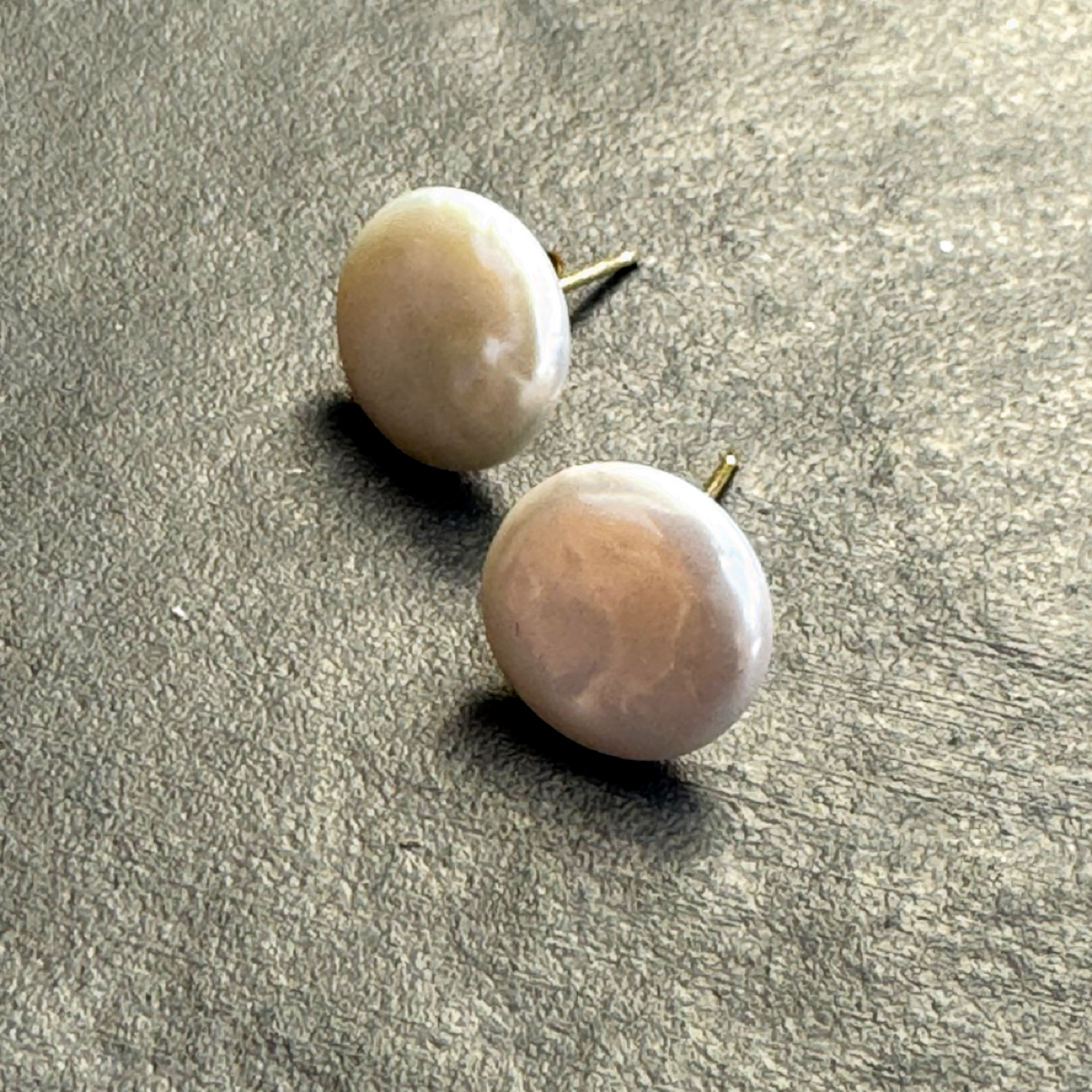 Two round pearl stud earrings with gold posts, resting on a textured gray surface.