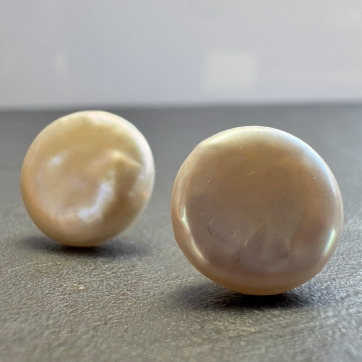 Two round, lustrous pearl stud earrings in a warm ivory-beige tone, photographed on a dark gray surface with a soft light gray background.