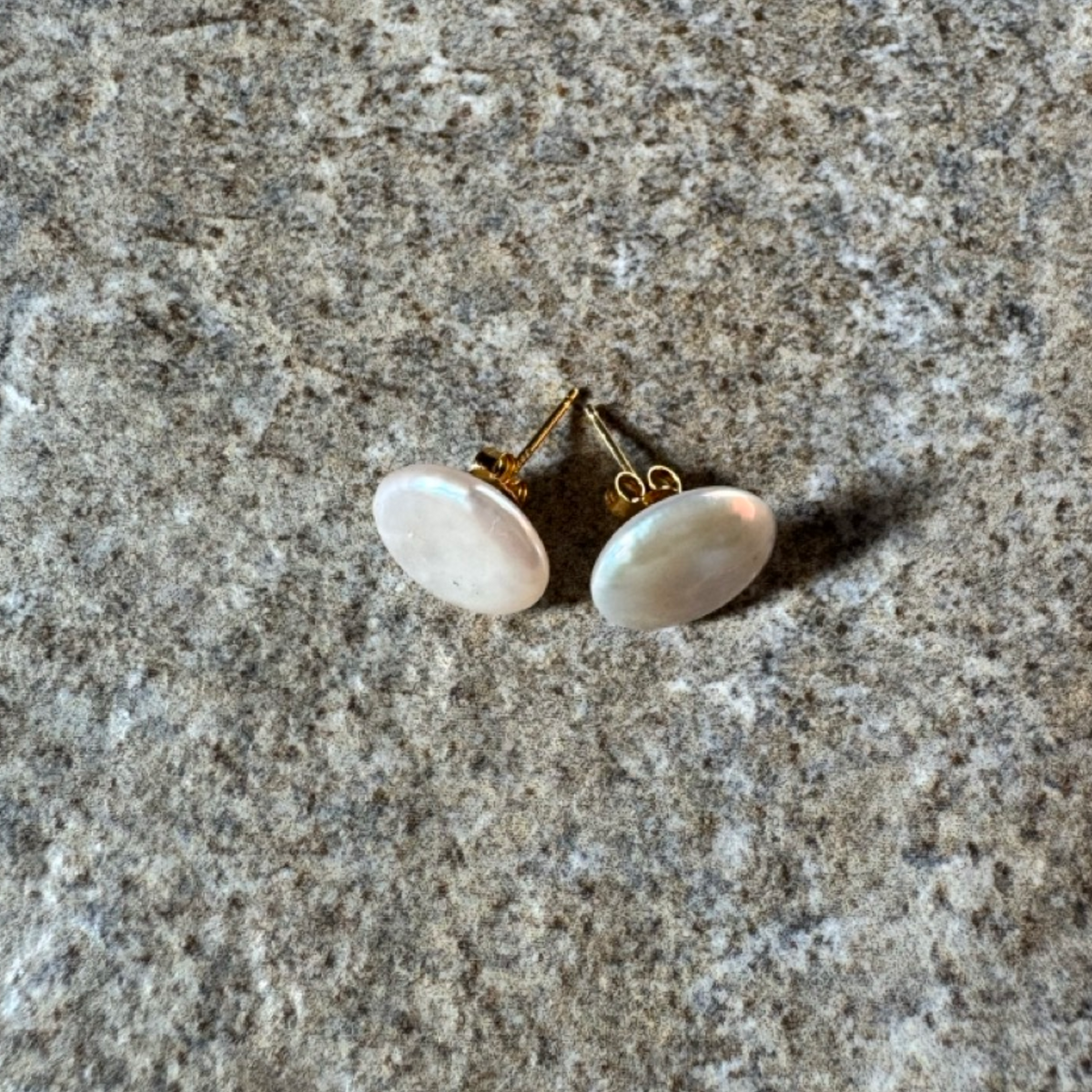 A pair of round stud earrings with iridescent white pearl coins and gold posts, resting on a textured gray stone surface.