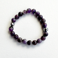 A beaded stretch bracelet made of polished purple and white marbled beads, with two small metallic silver spacer beads, displayed on a white textured fabric background.