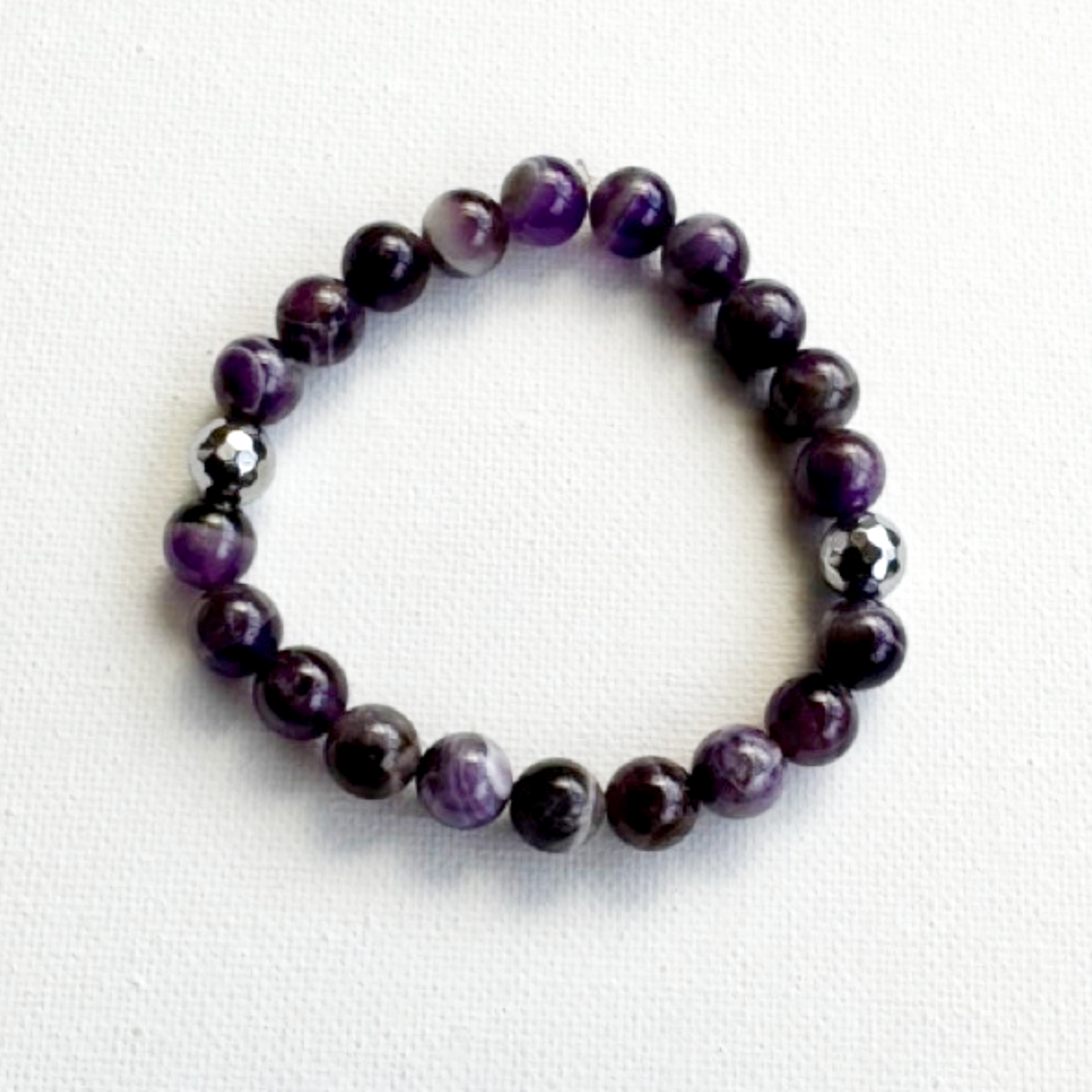 A beaded stretch bracelet made of polished purple and white marbled beads, with two small metallic silver spacer beads, displayed on a white textured fabric background.
