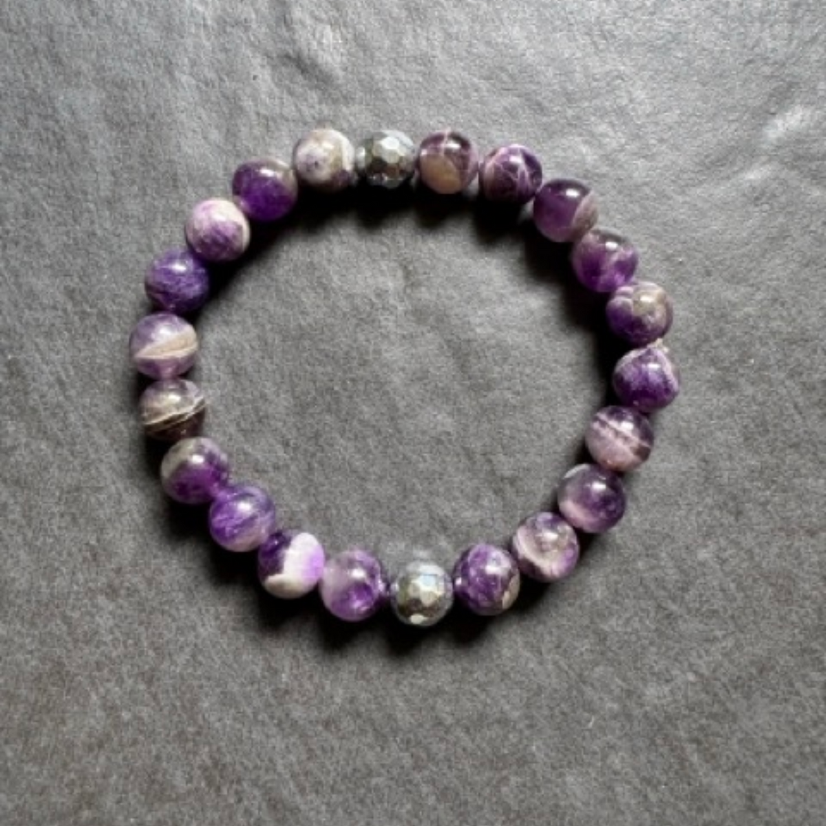A beaded stretch bracelet made of polished purple and white marbled gemstone beads, displayed on a textured gray slate surface.
