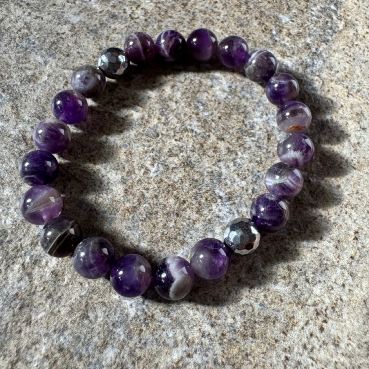 A beaded stretch bracelet made of polished purple and smoky gray gemstone beads, displayed on a textured gray stone surface.
