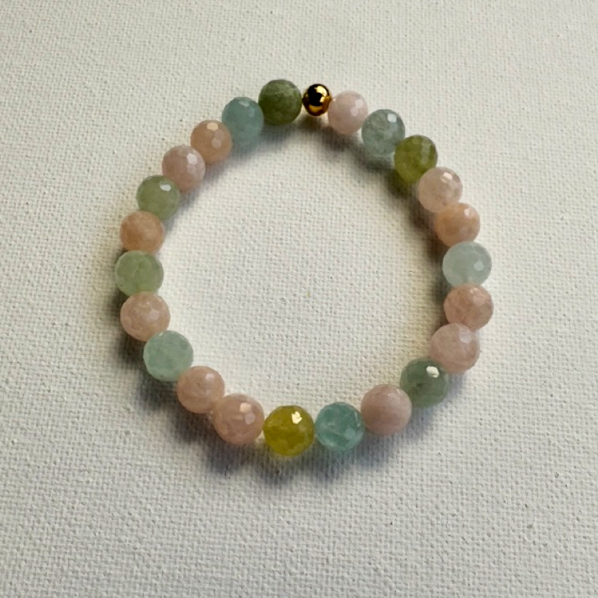 A beaded stretch bracelet made of faceted round beads in pastel pink, light green, and pale blue, with a small gold bead at the top, displayed on a textured off-white fabric background.