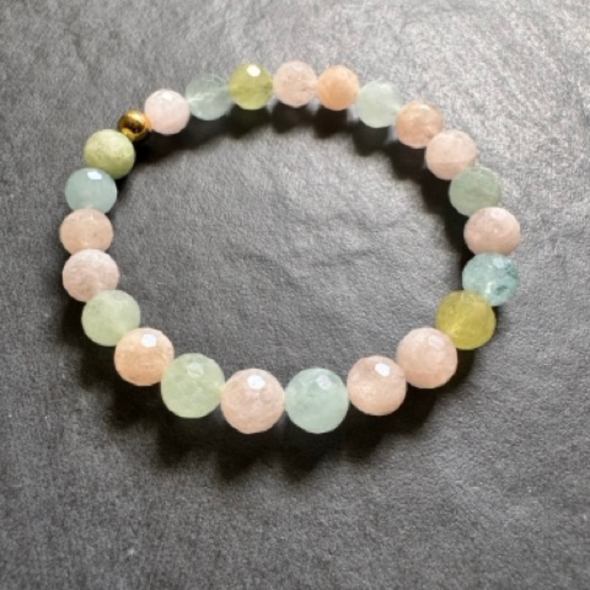 A beaded stretch bracelet made of faceted round gemstone beads in pastel pink, light green, and pale yellow, with a small gold spacer bead, displayed on a dark gray textured surface.