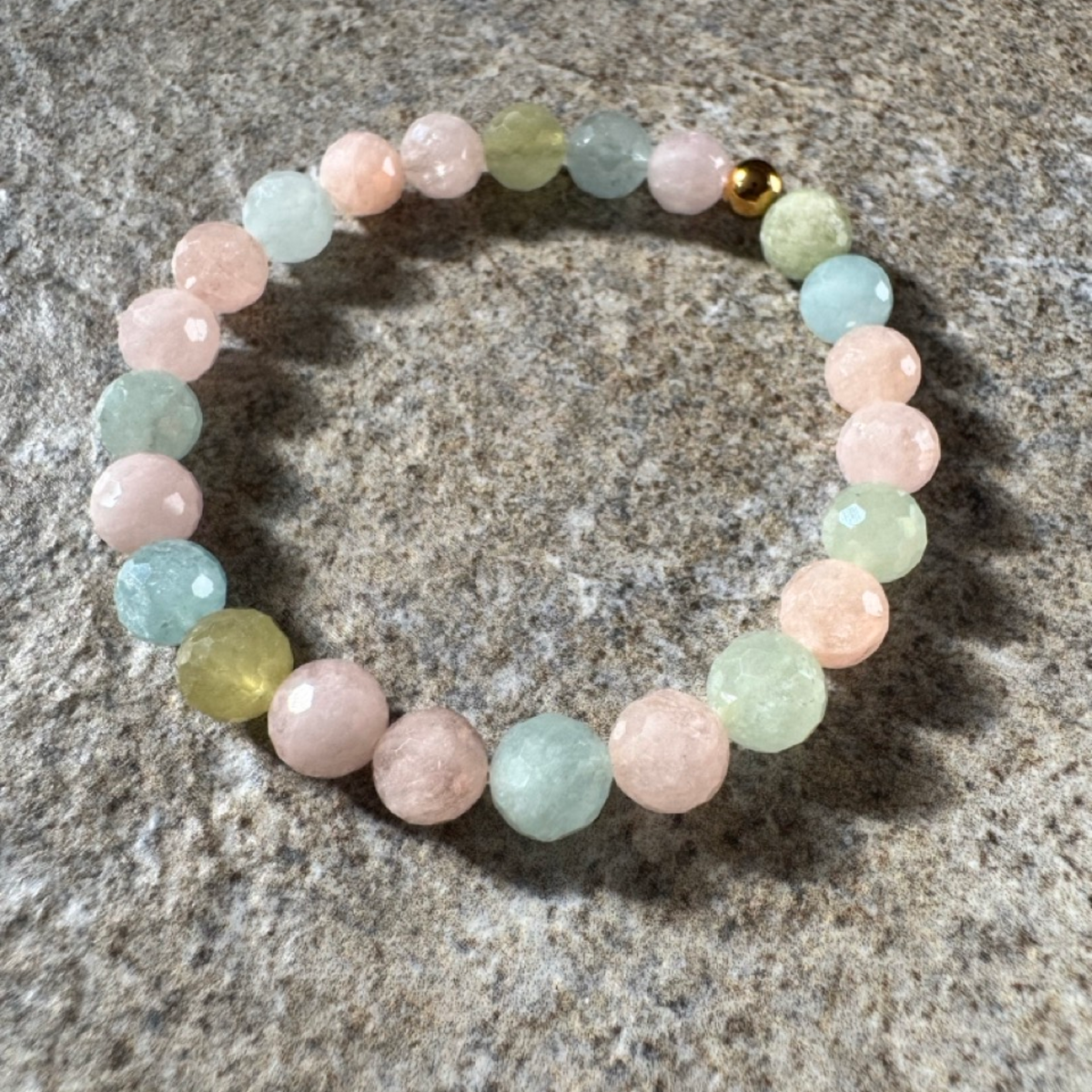 A beaded stretch bracelet made of faceted pastel gemstone beads in pink, light green, and light blue, with a small gold accent bead, displayed on a textured gray stone surface.