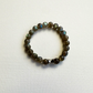 A beaded stretch bracelet made of labradorite beads with a shimmering green-blue iridescent finish, featuring a polished silver-toned metal focal, displayed on a textured off-white fabric background.